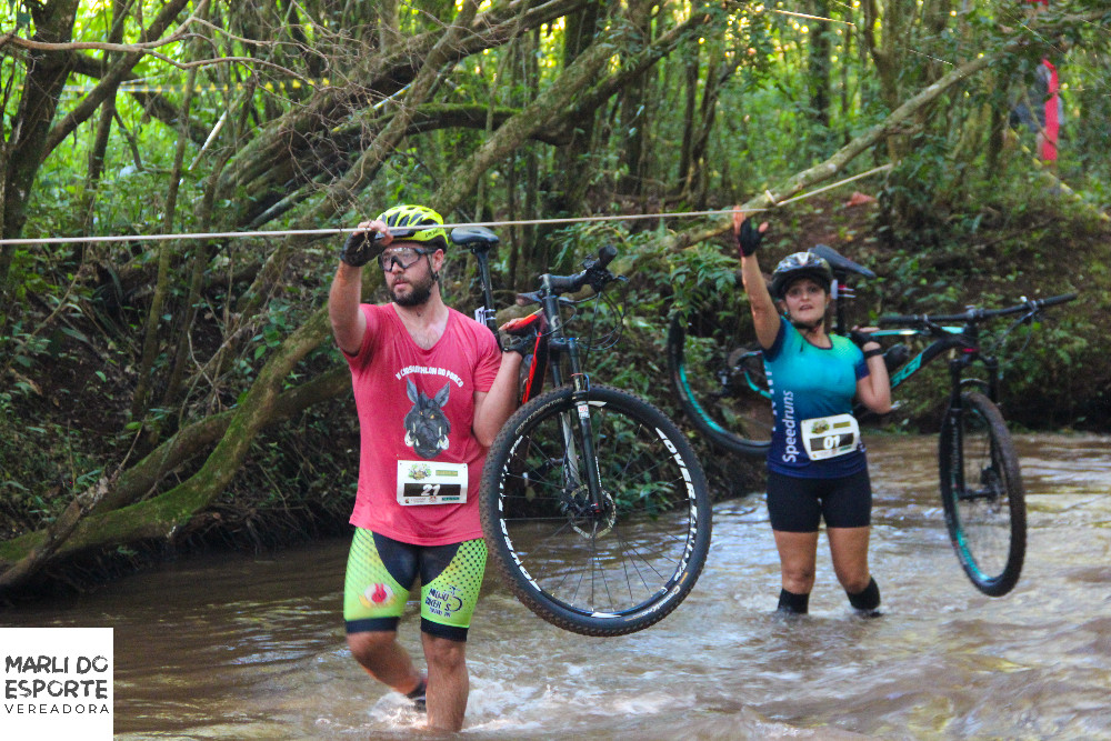 #Pracegover: Fotografia. Dois atletas, um homem e uma mulher, atravessa, um córrego segundo suas bicicletas com a mão esquerda, e com a mão direita seguram em uma corda suspensa.