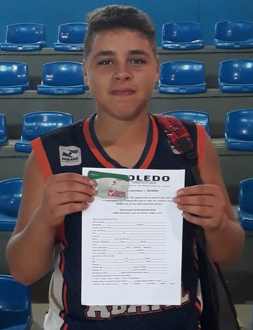 #Pracegover. Fotografia. Um adolescente posa em frente as arquibancadas de um ginásio de esportes. Na sua mão o cartão do passe atleta e o termo de compromisso.