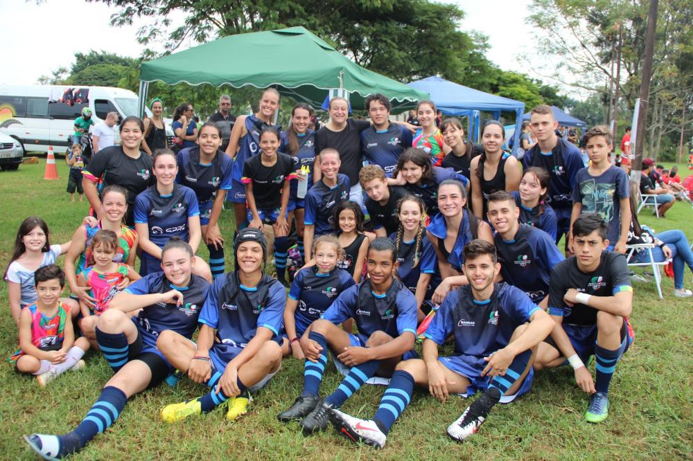 #Pracegover: Fotografia. Vários atletas de rugby, homens e mulheres posando para a foto, alguns sentados, outros em pé. Usam uniforme azul, ao fundo uma tenda verde.