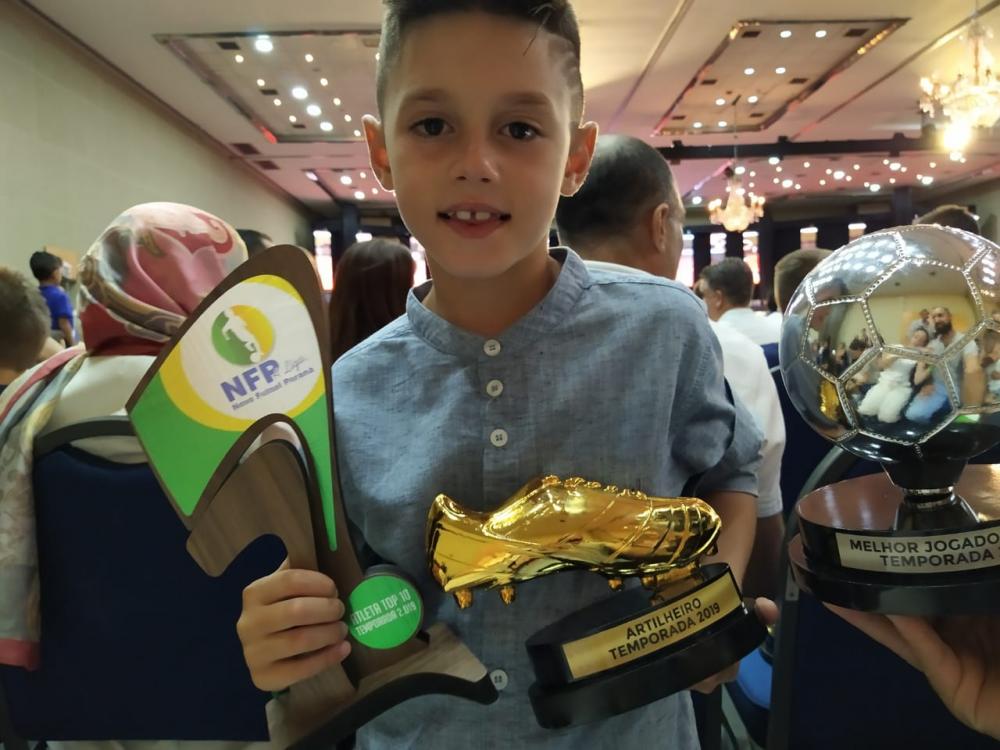 #Pracegover: Fotografia. Davi veste camisa cinza e em suas mãos segura os troféus da premiação.