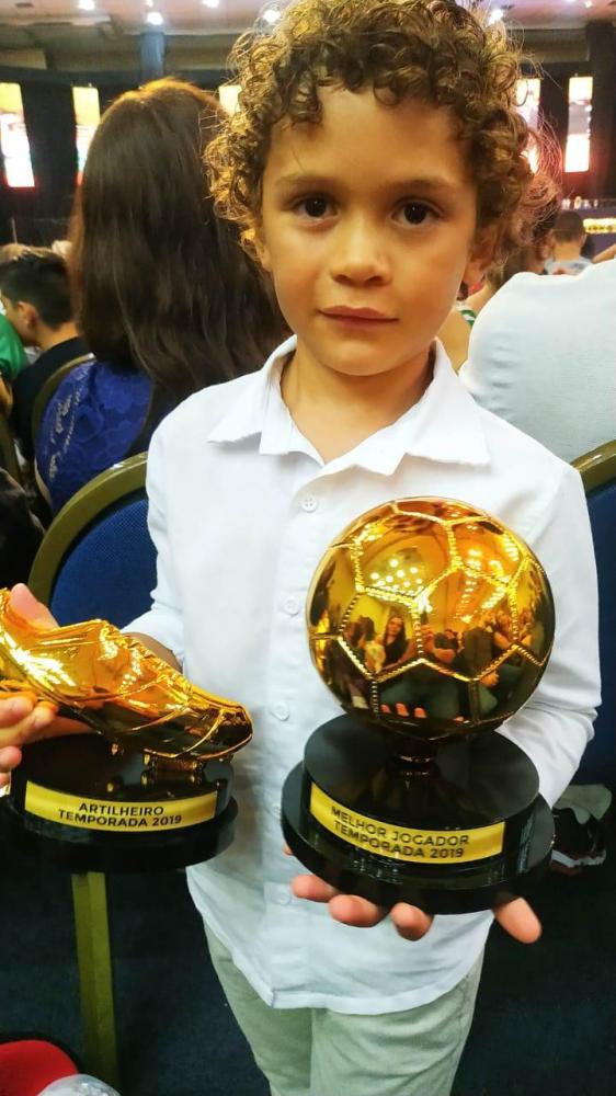 #Pracegover: Fotografia. Enzo veste camisa branca, e, segura em suas mãos dois troféus.